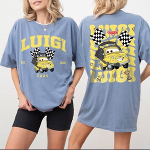 Peut inclure: T-shirt bleu clair avec un dessin de voiture de dessin anim&eacute; jaune, des drapeaux &agrave; damiers et le nom "Luigi". Le dos du t-shirt r&eacute;p&egrave;te le graphisme de la voiture et le nom "Luigi". Le t-shirt porte &eacute;galement le texte "EST. 2006" et "Route 66".