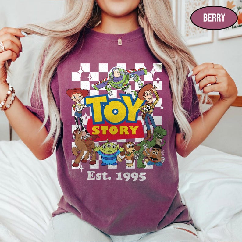 May include: Berry-colored t-shirt featuring a graphic of Toy Story characters, including Woody, Buzz Lightyear, and others. The words "TOY STORY" are in yellow and blue, with "Est. 1995" below. The design is set against a checkered background.