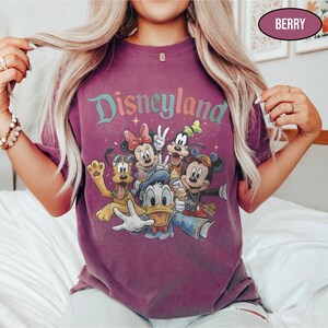 May include: Berry-colored t-shirt featuring the word "Disneyland" in a colorful, whimsical font. The shirt showcases a graphic of Disney characters including Mickey Mouse, Minnie Mouse, Donald Duck, Goofy, and Pluto. The shirt has a vintage, faded look.