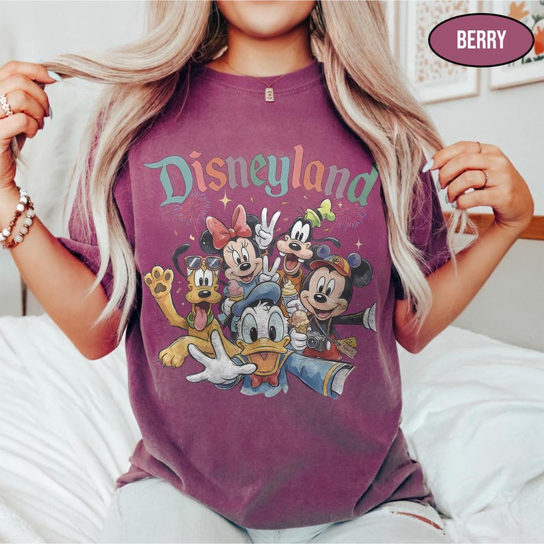 May include: Berry-colored t-shirt featuring the word "Disneyland" in a colorful, whimsical font. The shirt showcases a graphic of Disney characters including Mickey Mouse, Minnie Mouse, Donald Duck, Goofy, and Pluto. The shirt has a vintage, faded look.