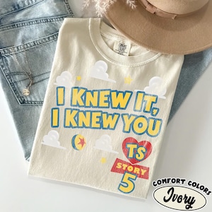 Puede incluir: Una camiseta de color marfil con el texto azul y amarillo I KNEW IT, I KNEW YOU, acompa&ntilde;ada de gr&aacute;ficos de nubes blancas, una estrella amarilla, una pelota colorida y un coraz&oacute;n rojo con las letras TS sobre el texto rojo STORY 5 en azul.