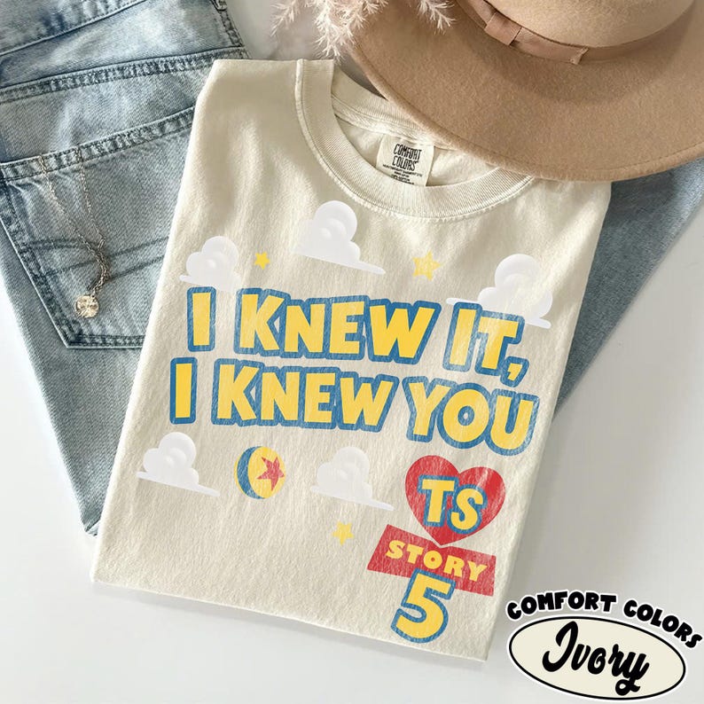 Puede incluir: Una camiseta de color marfil con el texto azul y amarillo I KNEW IT, I KNEW YOU, acompa&ntilde;ada de gr&aacute;ficos de nubes blancas, una estrella amarilla, una pelota colorida y un coraz&oacute;n rojo con las letras TS sobre el texto rojo STORY 5 en azul.