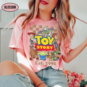 May include: Pink t-shirt featuring a colorful graphic of Toy Story characters with the text "TOY STORY" and "Est. 1995". The design includes Woody, Buzz Lightyear, and other characters from the animated film. The shirt has a crew neck.
