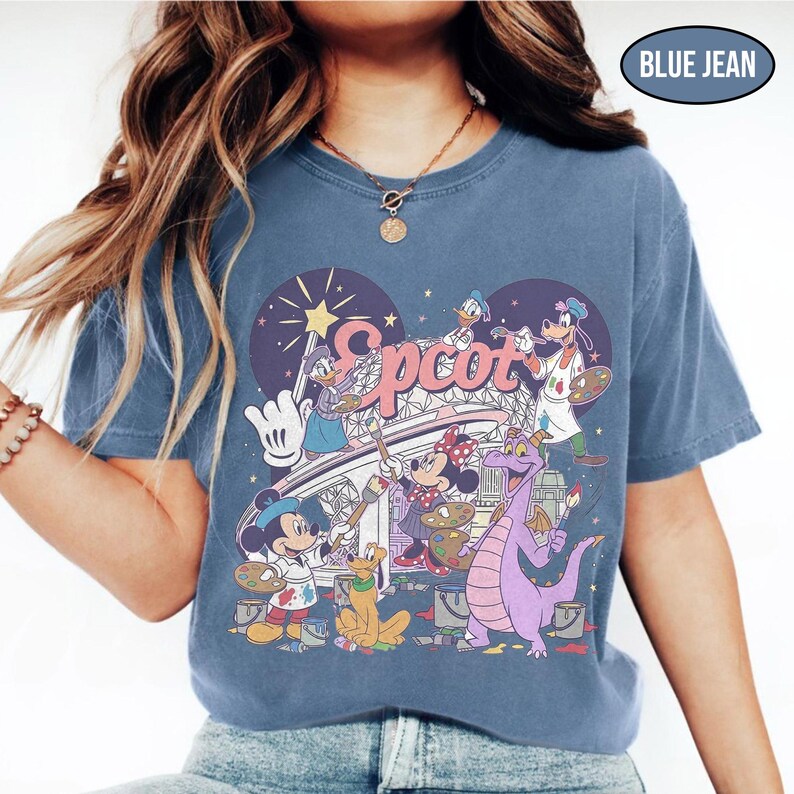 May include: Blue jean colored t-shirt featuring a colorful graphic of Disney characters painting the word "Epcot". The design includes Mickey Mouse, Minnie Mouse, Donald Duck, Goofy, and Figment. The shirt has a crew neck and short sleeves.