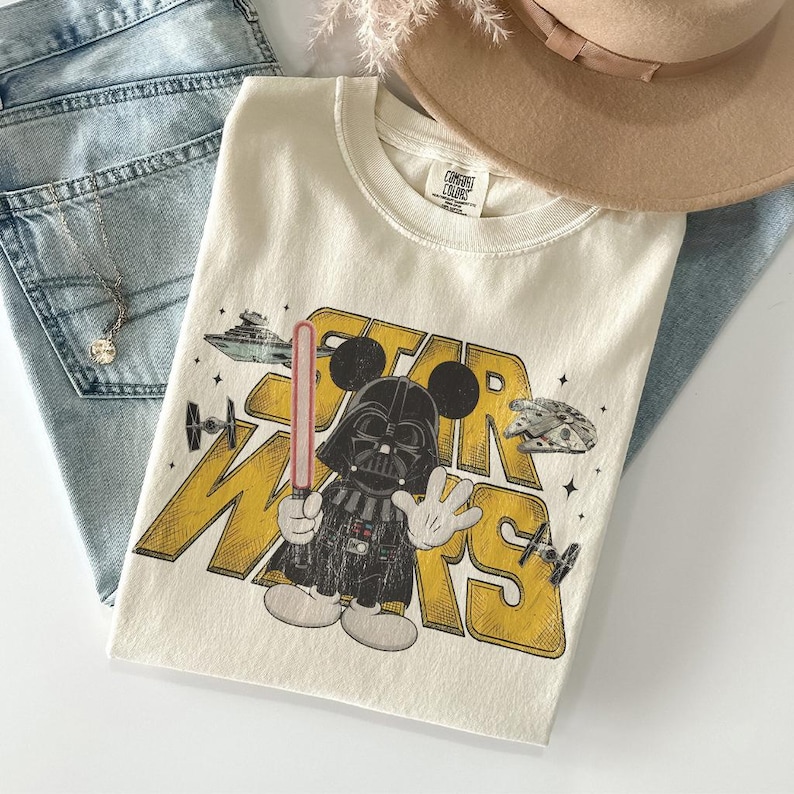 Puede incluir: Camiseta color crema con un gr&aacute;fico de Darth Vader como Mickey Mouse con las palabras "STAR WARS" en amarillo. La camiseta incluye im&aacute;genes de naves espaciales y un sable de luz. La camiseta est&aacute; doblada sobre unos vaqueros azules y un sombrero beige.