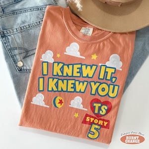 Puede incluir: Una camiseta de manga corta color naranja quemado con el texto "I KNEW IT, I KNEW YOU" en amarillo con bordes azules. El dise&ntilde;o incluye nubes blancas, una estrella amarilla, un coraz&oacute;n rojo con "TS" dentro y el texto "STORY 5" en amarillo.