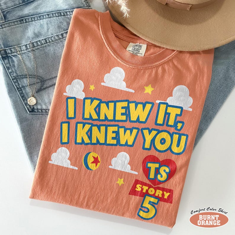 Puede incluir: Una camiseta de manga corta color naranja quemado con el texto "I KNEW IT, I KNEW YOU" en amarillo con bordes azules. El dise&ntilde;o incluye nubes blancas, una estrella amarilla, un coraz&oacute;n rojo con "TS" dentro y el texto "STORY 5" en amarillo.