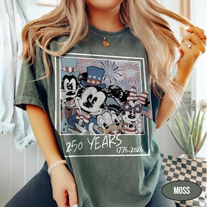 May include: A dark green t-shirt featuring a square graphic of cartoon characters wearing patriotic hats and star-shaped glasses with fireworks in the background. The text below the graphic reads 250 YEARS 1776-2026 in a white handwritten font.
