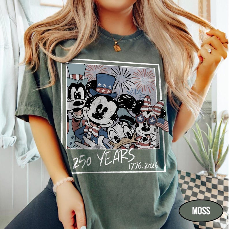 May include: A dark green t-shirt featuring a square graphic of cartoon characters wearing patriotic hats and star-shaped glasses with fireworks in the background. The text below the graphic reads 250 YEARS 1776-2026 in a white handwritten font.