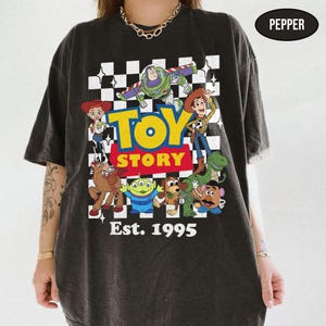 May include: Dark gray t-shirt featuring the Toy Story logo and characters, including Woody, Buzz Lightyear, and others, against a checkered background. The text "Est. 1995" is printed below the logo.