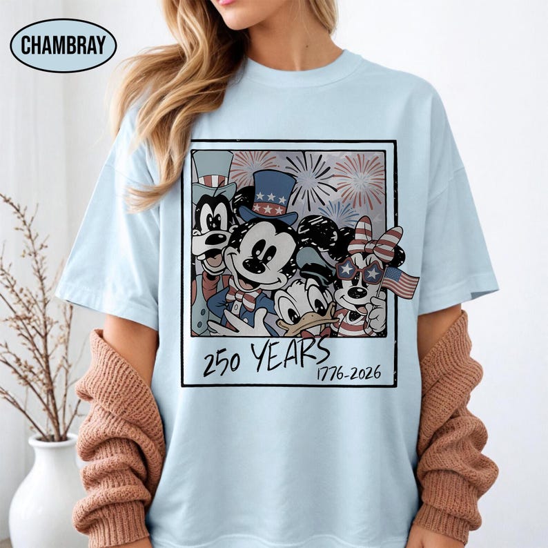 May include: A light blue t-shirt featuring a rectangular graphic of cartoon characters wearing patriotic hats and accessories. The design includes fireworks, a small American flag, and the text 250 YEARS 1776-2026. The word CHAMBRAY is printed in a small oval at the top left.