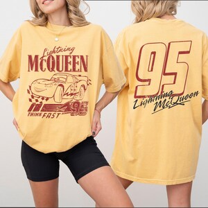 Puede incluir: Camiseta amarilla mostaza con gr&aacute;ficos de Lightning McQueen. El dise&ntilde;o frontal incluye el texto "Lightning McQueen", una ilustraci&oacute;n de coche de carreras y "95". La parte trasera muestra un gran "95" y "Lightning McQueen" en cursiva.
