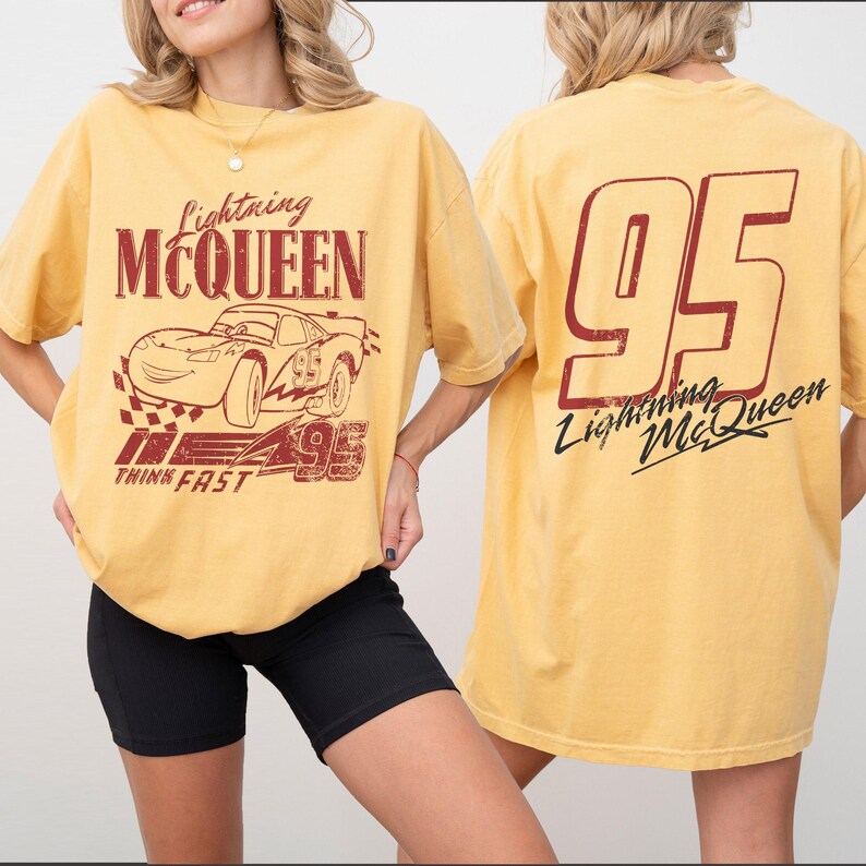 Puede incluir: Camiseta amarilla mostaza con gr&aacute;ficos de Lightning McQueen. El dise&ntilde;o frontal incluye el texto "Lightning McQueen", una ilustraci&oacute;n de coche de carreras y "95". La parte trasera muestra un gran "95" y "Lightning McQueen" en cursiva.