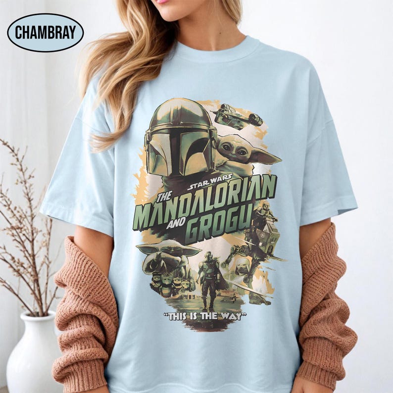 May include: Light blue t-shirt featuring a graphic from "The Mandalorian and Grogu." The design includes the Mandalorian's helmet, Grogu, and the text "The Mandalorian and Grogu" and "This is the Way."