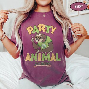 May include: Berry-colored t-shirt featuring a cartoon sloth wearing sunglasses and a green shirt, with the words "PARTY ANIMAL" in yellow and green. The shirt also has the text "ZOOTOPIA BETTER TOGETHER".
