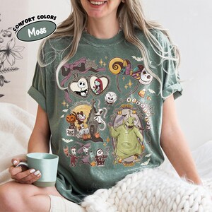 May include: A moss green t-shirt featuring a colorful graphic design from "The Nightmare Before Christmas." The design includes characters like Jack Skellington, Sally, and Oogie Boogie. The shirt has rolled-up sleeves and the words "Comfort Colors" and "Moss" are printed on the top left.