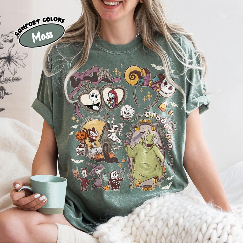 May include: A moss green t-shirt featuring a colorful graphic design from "The Nightmare Before Christmas." The design includes characters like Jack Skellington, Sally, and Oogie Boogie. The shirt has rolled-up sleeves and the words "Comfort Colors" and "Moss" are printed on the top left.