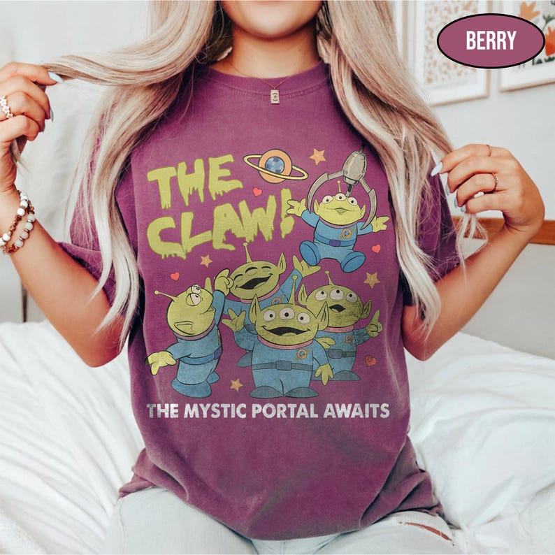 May include: Berry-colored t-shirt featuring a graphic of green aliens in blue uniforms with the text "THE CLAW!" and "THE MYSTIC PORTAL AWAITS" in yellow and white. The design includes a planet and stars.