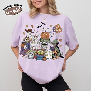 May include: A lilac Comfort Colors t-shirt with a Halloween cartoon graphic. The design features characters in costumes, including a pumpkin-headed robot, ghosts, and a dinosaur reading. The text "orchid" is in the upper left corner of the t-shirt.
