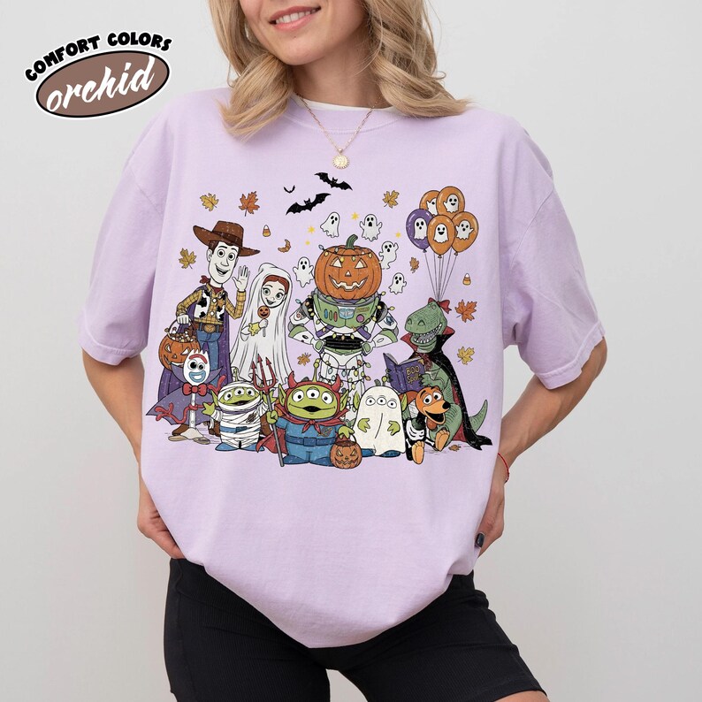 May include: A lilac Comfort Colors t-shirt with a Halloween cartoon graphic. The design features characters in costumes, including a pumpkin-headed robot, ghosts, and a dinosaur reading. The text "orchid" is in the upper left corner of the t-shirt.