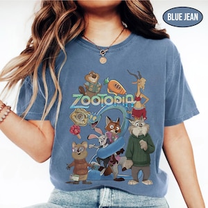 May include: A blue jean colored t-shirt featuring the word "Zootopia" in a stylized font, with cartoon characters from the movie. The shirt has a crew neck and short sleeves. The graphic includes a fox, rabbit, and other animal characters.