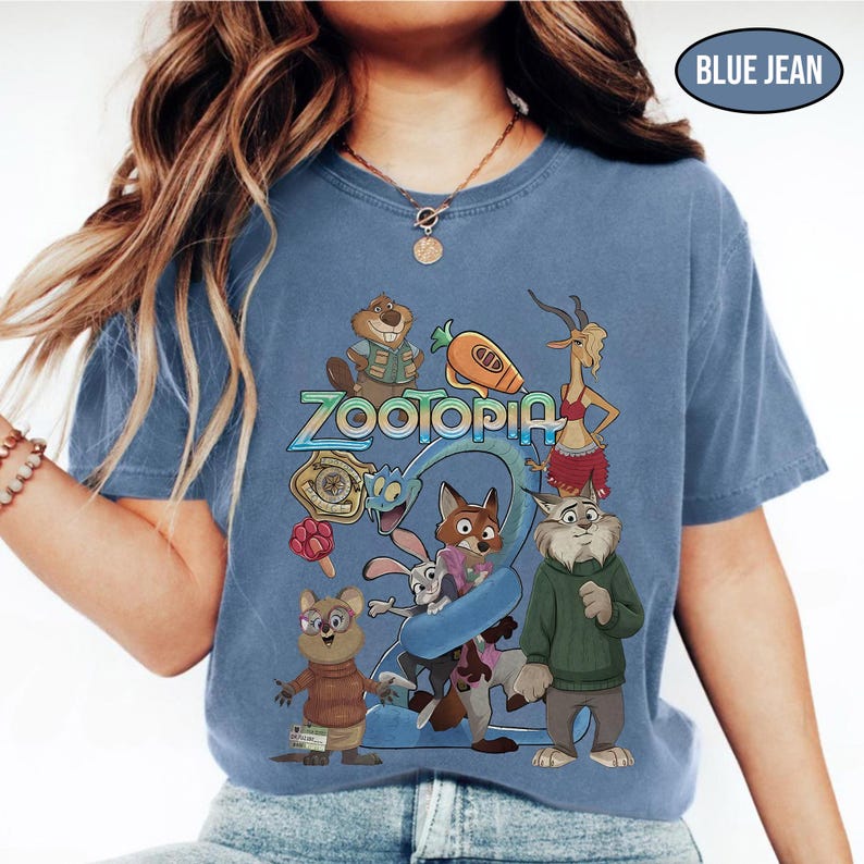 May include: A blue jean colored t-shirt featuring the word "Zootopia" in a stylized font, with cartoon characters from the movie. The shirt has a crew neck and short sleeves. The graphic includes a fox, rabbit, and other animal characters.