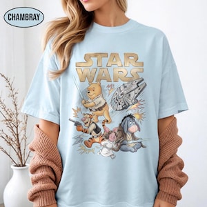 Puede incluir: Camiseta azul claro con un dise&ntilde;o que combina Star Wars y Winnie the Pooh. El texto dorado dice "STAR WARS" sobre ilustraciones de Pooh, Tigger, Piglet, Eeyore y el Halc&oacute;n Milenario. La camiseta tiene mangas cortas.