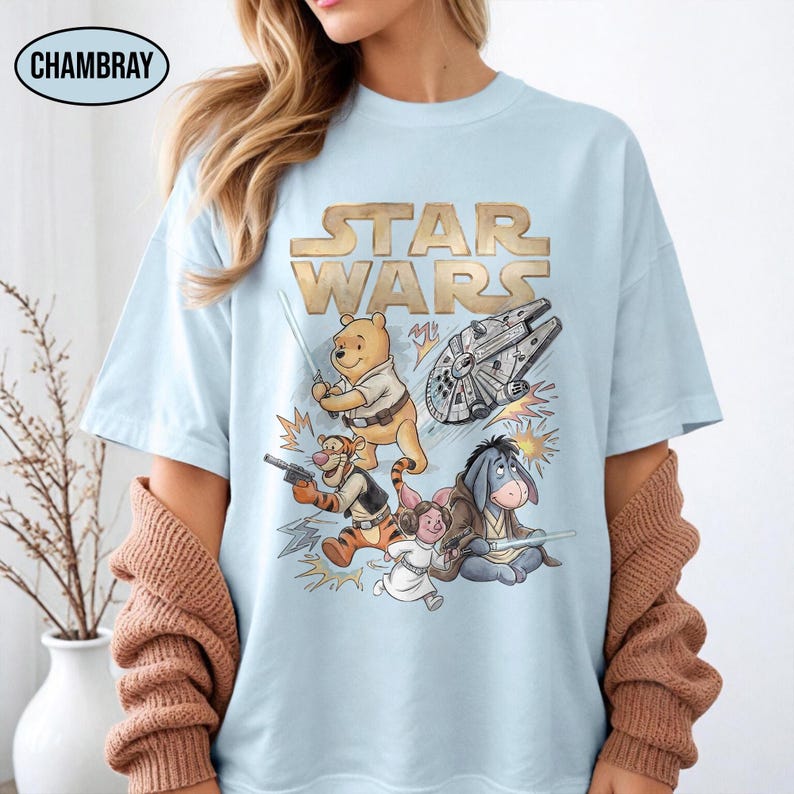 Puede incluir: Camiseta azul claro con un dise&ntilde;o que combina Star Wars y Winnie the Pooh. El texto dorado dice "STAR WARS" sobre ilustraciones de Pooh, Tigger, Piglet, Eeyore y el Halc&oacute;n Milenario. La camiseta tiene mangas cortas.