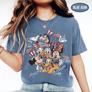 Może przedstawiać: Niebieska koszulka w kolorze dżinsu z grafiką postaci Disneya Myszki Miki, Myszki Minnie, Kaczora Donalda, Kaczki Daisy, Goofy'ego i Pluto, z amerykańskimi flagami i patriotycznymi kapeluszami. Koszulka upamiętnia 250. rocznicę USA 1776-2026.