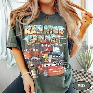May include: A dark green t-shirt featuring a graphic print of characters from the movie Cars. The design includes the text Radiator Springs and Lightning McQueen, along with a gold trophy, a traffic light, and checkered flags in the illustration.