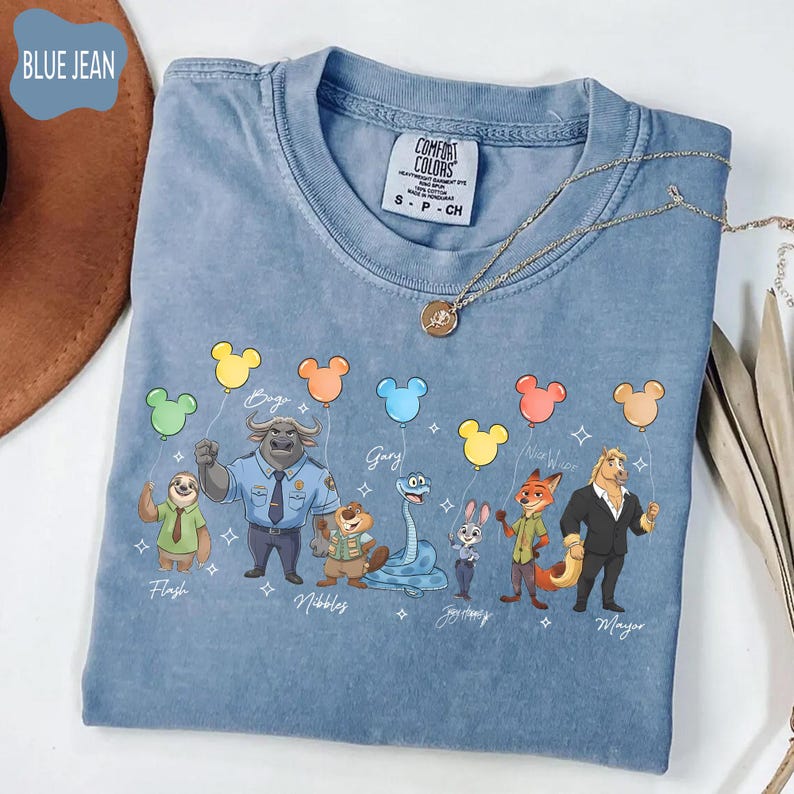 Puede incluir: Camiseta azul vaquero Comfort Colors con un gr&aacute;fico de dibujos animados de personajes, cada uno con un globo de Mickey Mouse. Los personajes incluyen un perezoso, un b&uacute;falo, una serpiente, un conejo, un zorro y un caballo. Un collar dorado est&aacute; sobre la camiseta.