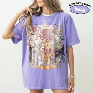 May include: A light purple t-shirt featuring a graphic of Winnie the Pooh characters in Halloween costumes. The print includes the text Disney Winnie the Pooh, Candy, and 100 Acre Wood. The design shows Pooh, Piglet, Eeyore, and Rabbit with pumpkins.
