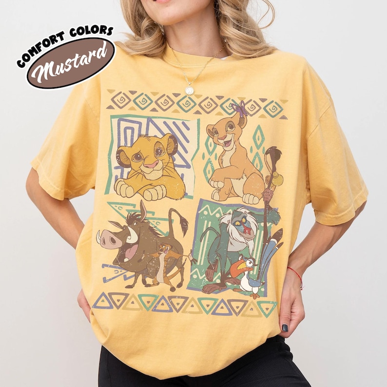 May include: A mustard yellow short-sleeve t-shirt featuring a graphic print of characters from The Lion King, including Simba, Nala, Pumbaa, Timon, Rafiki, and Zazu, arranged in four panels with geometric borders and the text Comfort Colors Mustard.