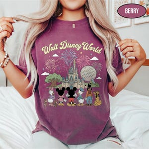 May include: Berry-coloured t-shirt with the text "Walt Disney World" above a graphic of Cinderella's castle, Spaceship Earth, and Disney characters. The design includes fireworks and a tree. The shirt is a short-sleeved crew neck.