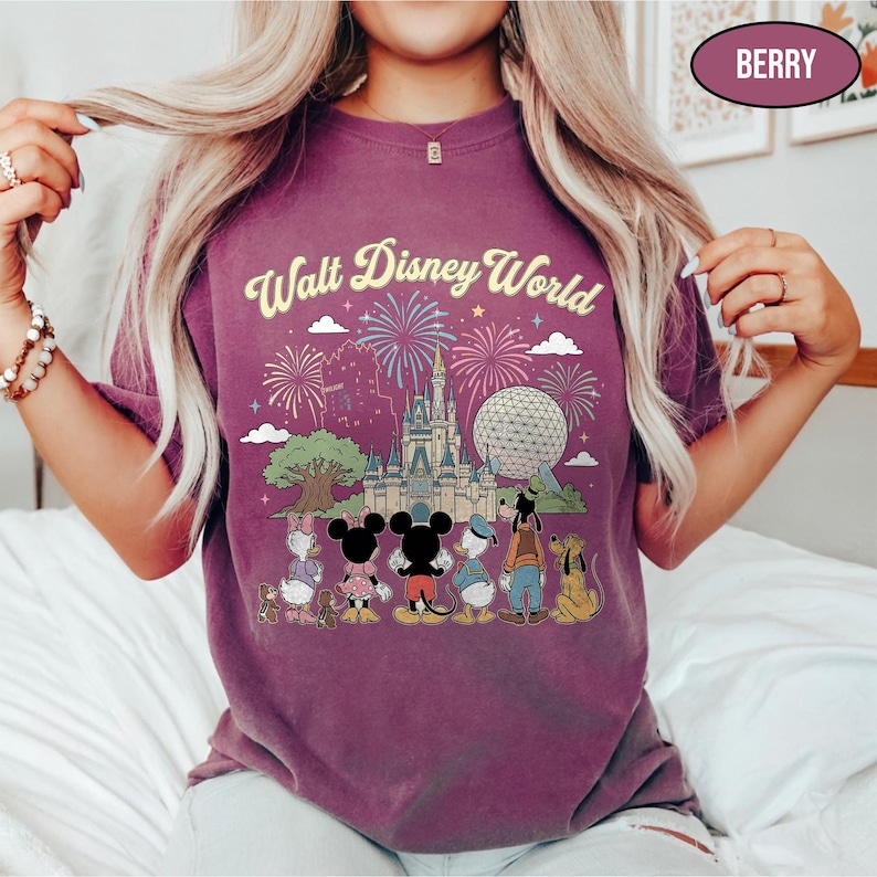 May include: Berry-coloured t-shirt with the text "Walt Disney World" above a graphic of Cinderella's castle, Spaceship Earth, and Disney characters. The design includes fireworks and a tree. The shirt is a short-sleeved crew neck.