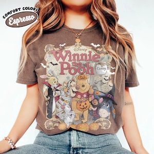 May include: A dark brown short-sleeve t-shirt featuring a graphic of Disney Winnie the Pooh characters dressed in Halloween costumes. The print includes the text Disney Winnie the Pooh, a candy jar, and pumpkins. The shirt has a Comfort Colors Espresso label.
