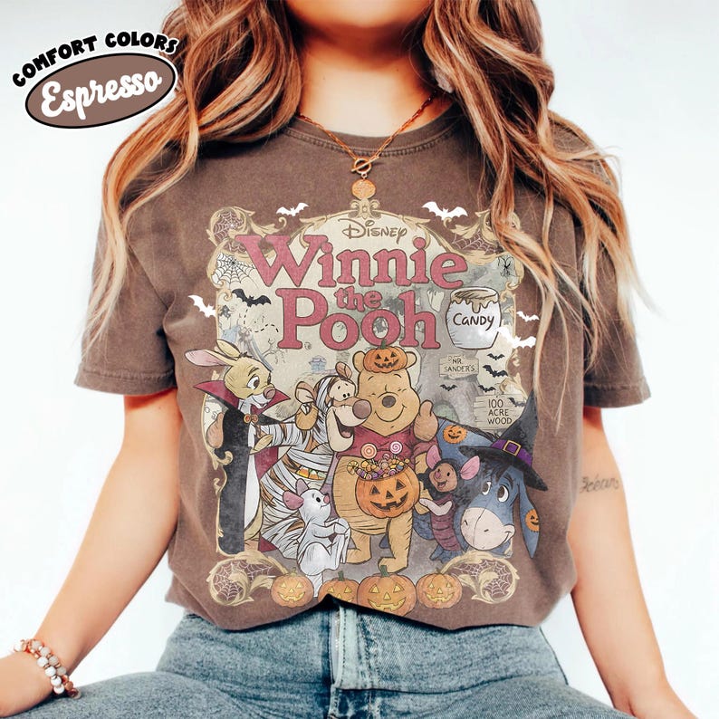 May include: A dark brown short-sleeve t-shirt featuring a graphic of Disney Winnie the Pooh characters dressed in Halloween costumes. The print includes the text Disney Winnie the Pooh, a candy jar, and pumpkins. The shirt has a Comfort Colors Espresso label.