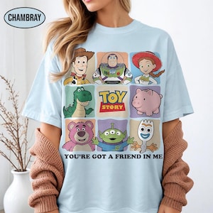 May include: Light blue t-shirt featuring a grid of Toy Story characters, including Woody, Buzz Lightyear, and Jessie. The shirt has the text "TOY STORY" and "YOU'RE GOT A FRIEND IN ME" printed on it. The shirt is made of a soft material.