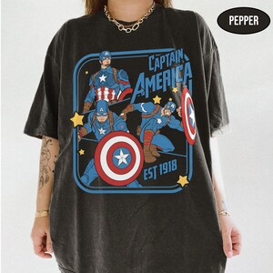 Może przedstawiać: Ciemnoszara koszulka z grafiką Kapitana Ameryki. Projekt zawiera wizerunki Kapitana Ameryki, jego tarczy oraz napis "Captain America EST 1918" w kolorze niebieskim i białym. Koszulka ma wygląd vintage.