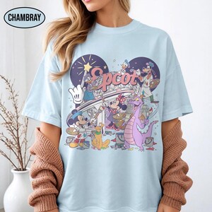 May include: Light blue t-shirt featuring a colorful graphic of Disney characters painting the word "Epcot" and a stylized structure. The design includes Mickey Mouse, Minnie Mouse, Donald Duck, Goofy, and Figment. The shirt has a crew neck and short sleeves.