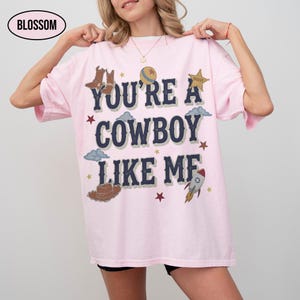 Puede incluir: Una camiseta de manga corta de color rosa claro con el texto azul oscuro "YOU'RE A COWBOY LIKE ME" rodeado de ilustraciones gr&aacute;ficas de botas de vaquero, una estrella de sheriff, un cohete, un sombrero, nubes y estrellas. La palabra "BLOSSOM" aparece en un &oacute;valo.