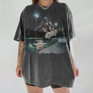 Pu&ograve; includere: Una t-shirt oversize grigio antracite con una stampa grafica raffigurante un personaggio mandaloriano che esegue un trick con lo skateboard in uno skatepark, con una piccola creatura verde su uno skateboard sottostante.