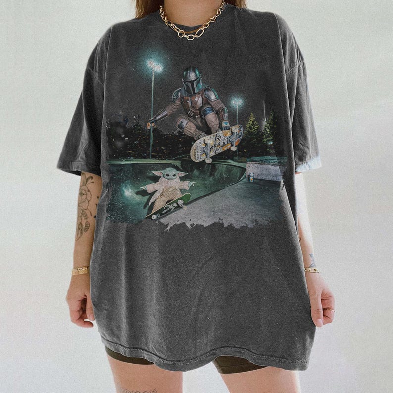 Pu&ograve; includere: Una t-shirt oversize grigio antracite con una stampa grafica raffigurante un personaggio mandaloriano che esegue un trick con lo skateboard in uno skatepark, con una piccola creatura verde su uno skateboard sottostante.
