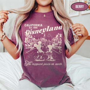 May include: Berry-colored t-shirt featuring a Disneyland graphic. The design includes the text "CALIFORNIA Est. 1955 Disneyland" with Mickey and Minnie Mouse. The phrase "The happiest place on earth" is also printed on the shirt.