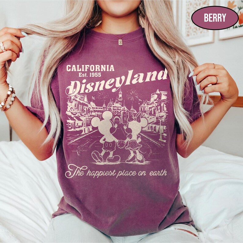 May include: Berry-colored t-shirt featuring a Disneyland graphic. The design includes the text "CALIFORNIA Est. 1955 Disneyland" with Mickey and Minnie Mouse. The phrase "The happiest place on earth" is also printed on the shirt.