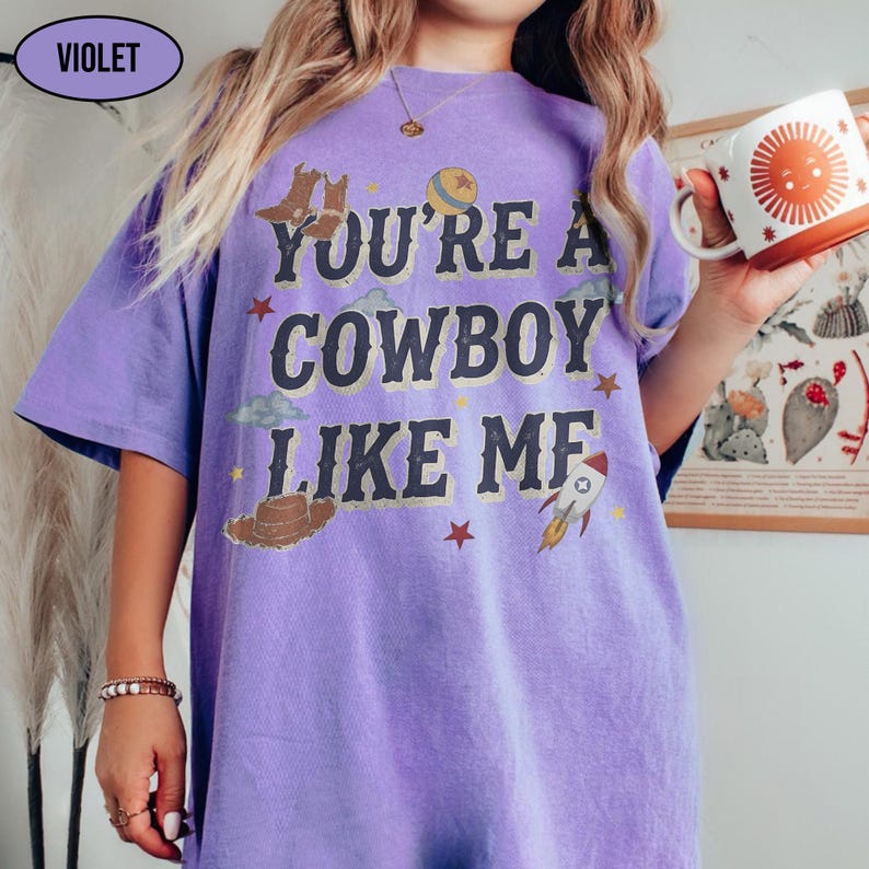 Puede incluir: Una camiseta violeta de manga corta con el texto "YOU'RE A COWBOY LIKE ME" en letras oscuras. El dise&ntilde;o incluye ilustraciones de botas de vaquero, una pelota, nubes, estrellas, un sombrero de vaquero y un cohete alrededor del texto.