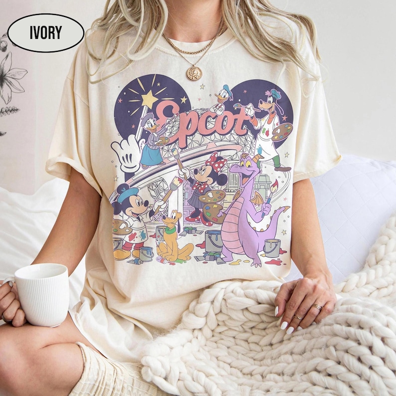 May include: Ivory-colored t-shirt featuring a colorful graphic of Disney characters painting the word "Epcot" in a whimsical style. The design includes Mickey Mouse, Minnie Mouse, Donald Duck, and a purple dragon. The shirt has short sleeves.