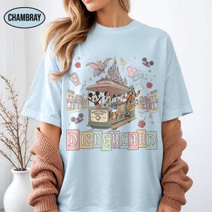 May include: Light blue t-shirt featuring a Disneyland design. The graphic includes Mickey Mouse, Minnie Mouse, Donald Duck, Goofy, and other characters riding a Main Street Trolley. The word "Disneyland" is displayed in colorful letters.