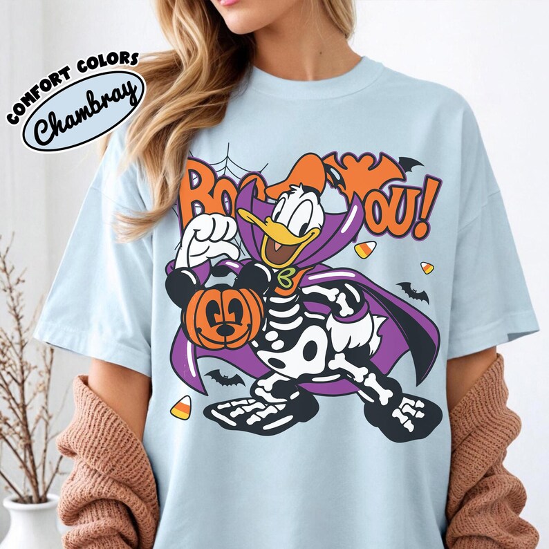 May include: A light blue t-shirt featuring a graphic of Donald Duck dressed in a skeleton costume and a purple cape, holding a Mickey Mouse-shaped pumpkin. The text "BOO TO YOU!" is printed above in orange, accompanied by bats, candy corn, and a spiderweb.