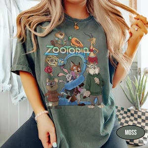May include: A moss green t-shirt featuring the word "Zootopia" in blue and yellow, with cartoon characters from the movie. The graphic includes a fox, rabbit, and other animals. The shirt is a comfortable, casual style.